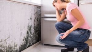 A woman crouching down in front of a wall with black mold.