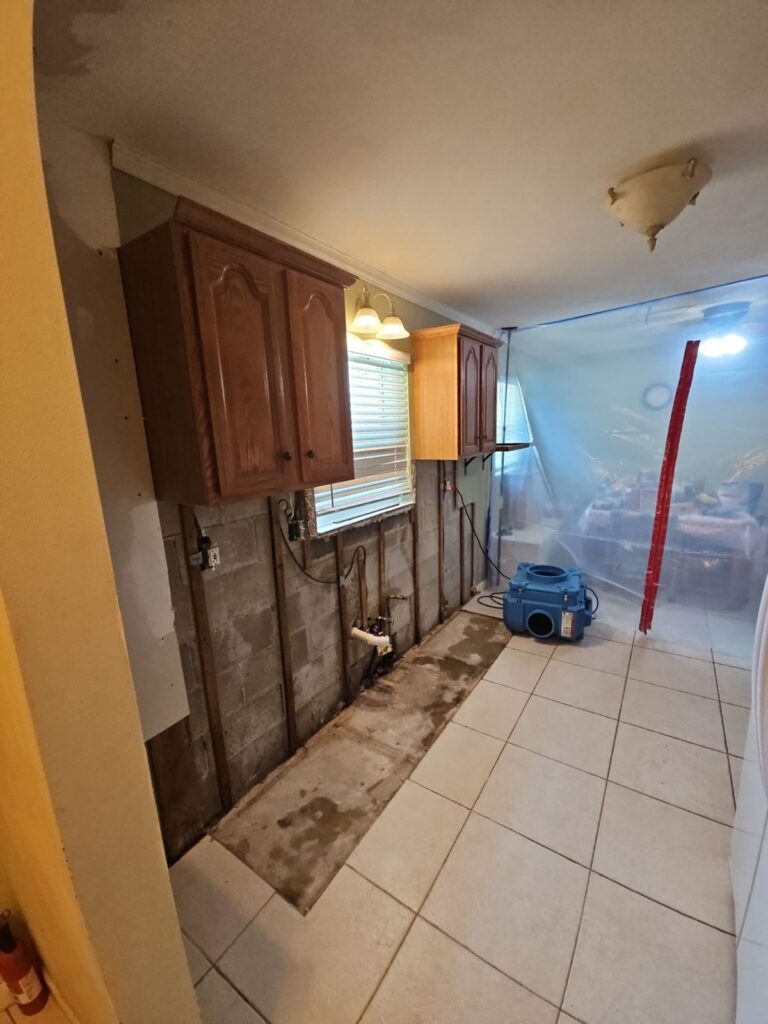 Titusville kitchen with lower cabinets and bottom drywall removed, containment plastic across the room and an air mover running